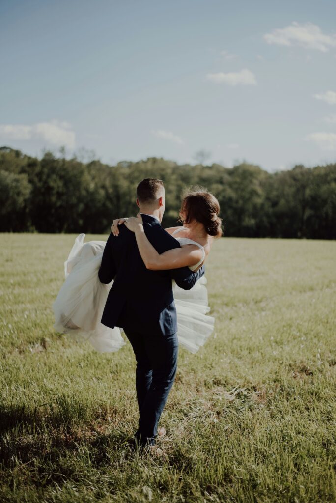Groom carrying his bride through a sunlit field, shot by a wedding photographer.