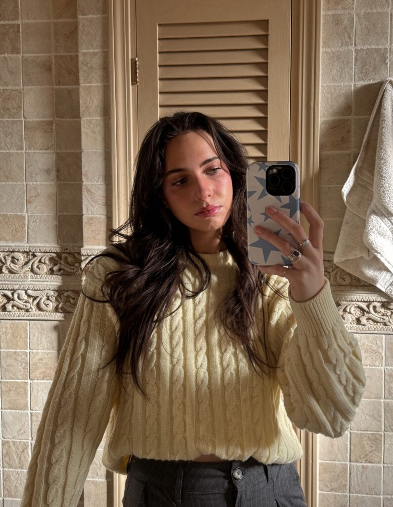 A woman creating for social media in a cream cable-knit sweater takes a mirror selfie in a warmly lit bathroom, holding her phone with a patterned case.