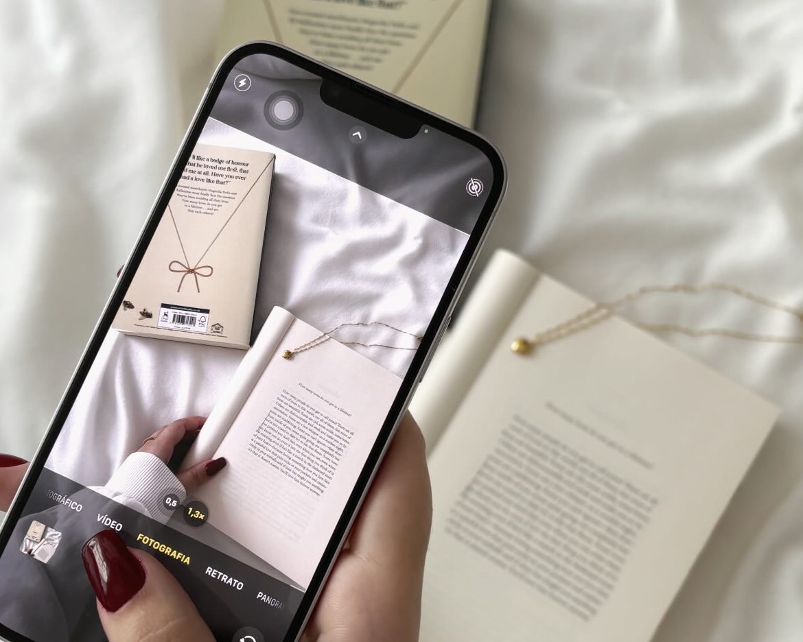 A close-up of a person taking a photo of open books and a gold necklace on white bedding, capturing an aesthetic flat-lay for social media content.