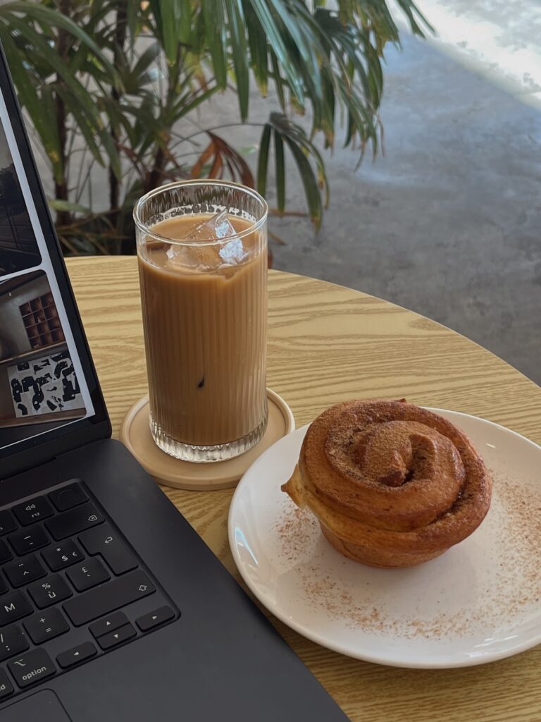 A cozy café scene with an open laptop, an iced coffee in a ribbed glass, and a cinnamon pastry on a white plate, set on a light wood table with plants in the background — a quiet moment from a blogging workflow.