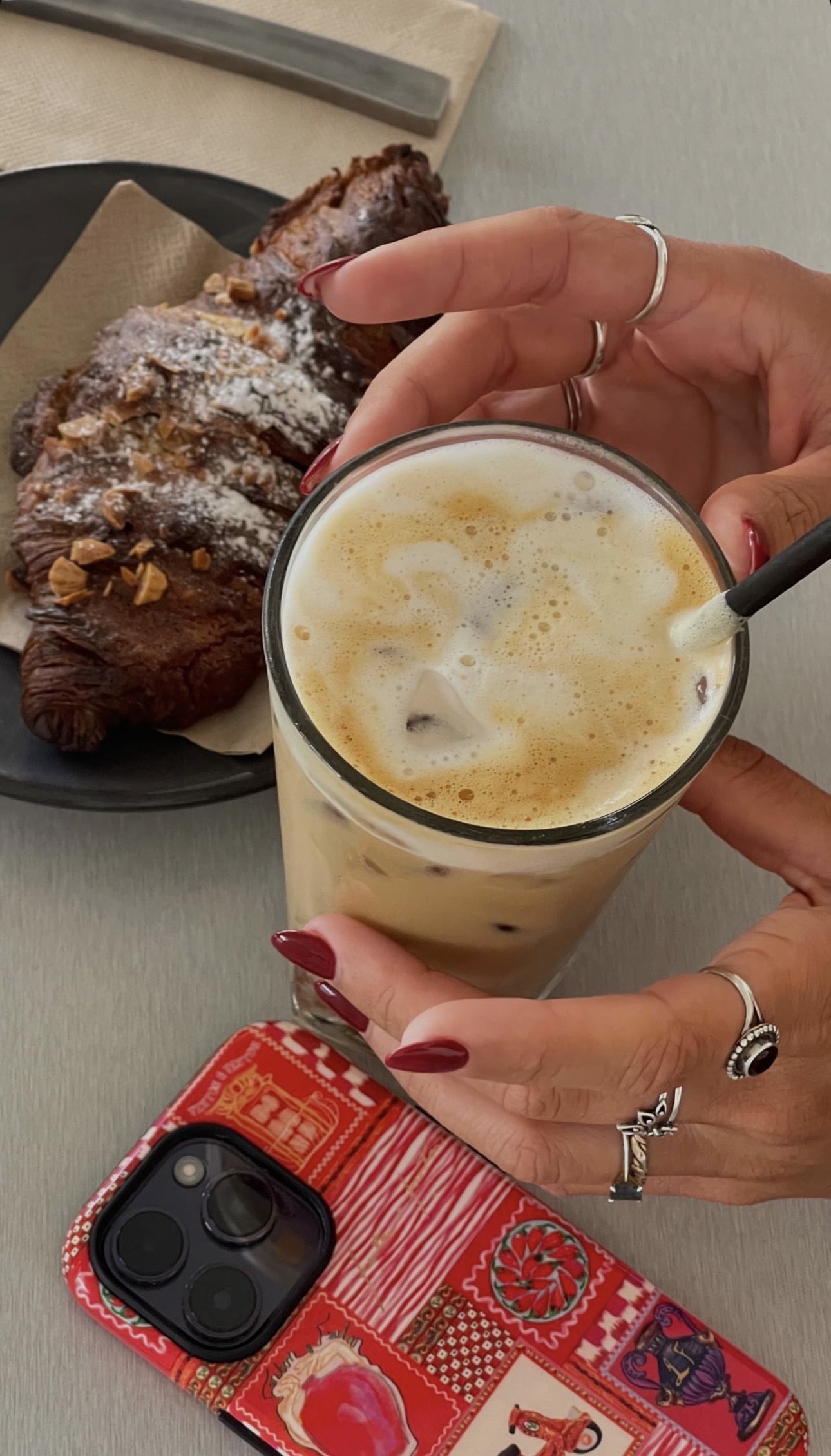A close-up café scene with hands holding an iced coffee, a chocolate croissant on a plate, and a red patterned phone case — a quiet behind-the-scenes moment from a blogging workday.