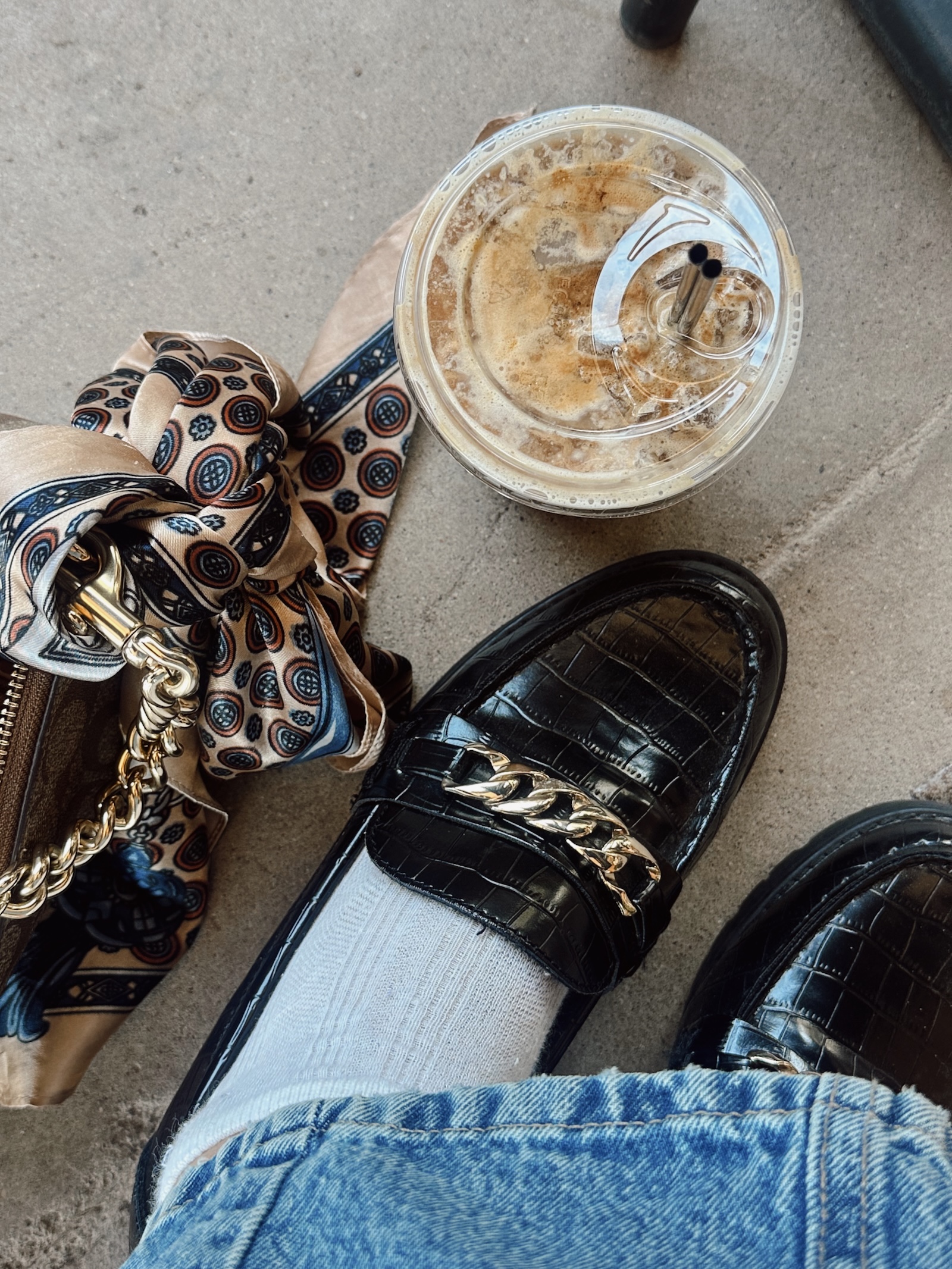 Overhead lifestyle photo of black crocodile loafers with gold chain detail, white socks, light denim, and an iced coffee in a plastic cup, representing long term content marketing and how to make content last longer through intentional, everyday creative workflows.