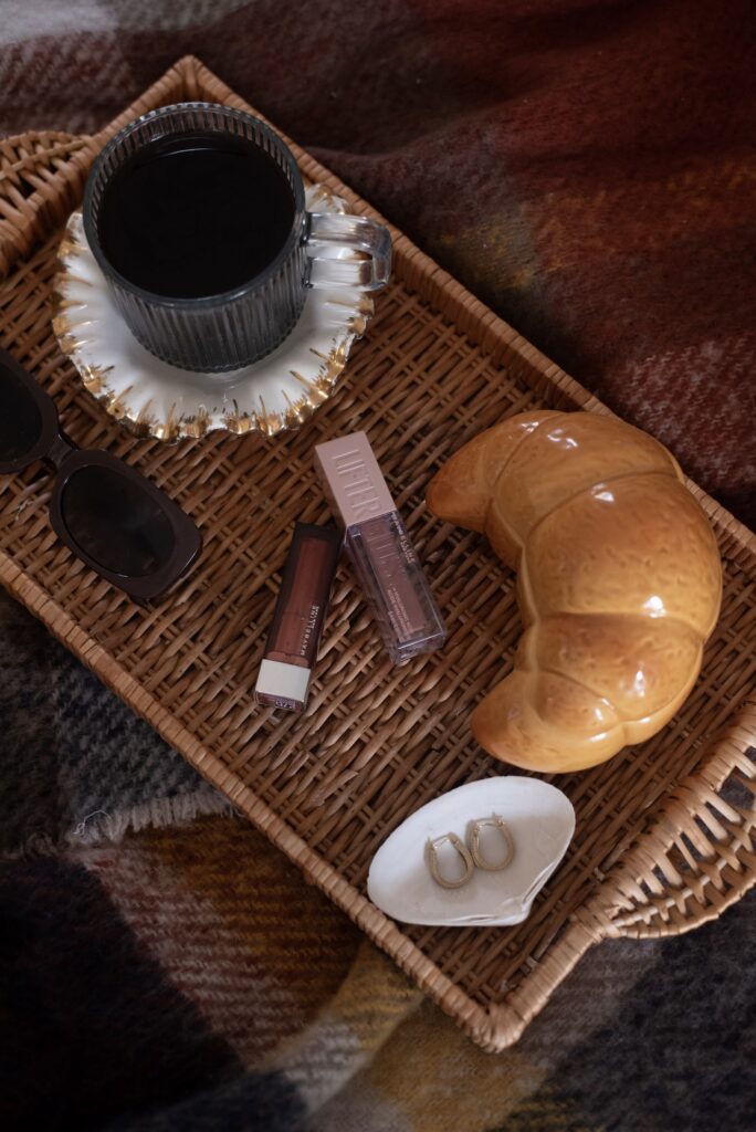 An aesthetic flatlay on a woven tray featuring a coffee cup, croissant, sunglasses, and makeup products—representing the "let it be fun" approach to Pinterest marketing and creative experimentation phase for new accounts.